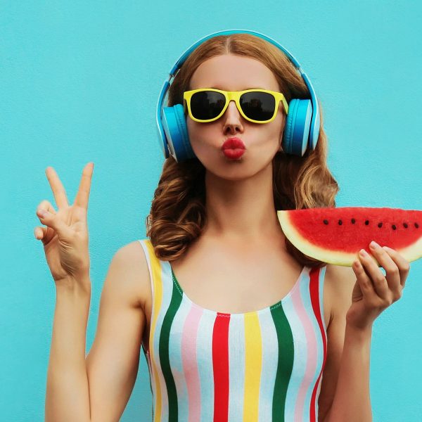 Summer fashion portrait of young woman in headphones listening to music with juicy slice of watermelon, female model blowing her lips posing on a colorful blue background